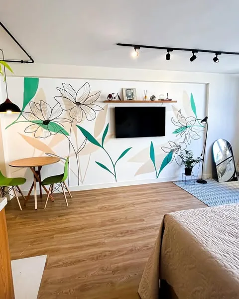 A mesma sala com mural floral em contornos pretos, folhas verdes e flores brancas ao redor da TV